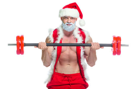 Christmas. Strong muscular Santa Claus with grey beard wearing christmas hat and red shorts is lifting a heavy barbell in a gym performing a workout isolated on white background.の写真素材
