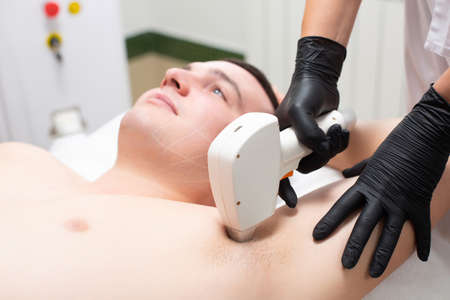 A young handsome man is lying on the couch of a cosmetology office with his hands raised during a laser hair removal procedure in the armpitの写真素材