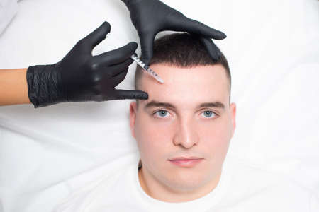 Close up of a young man face and the hands of a cosmetologist in black medical gloves injecting botox from wrinkles into the places marked with a white pencil on the patient foreheadの写真素材