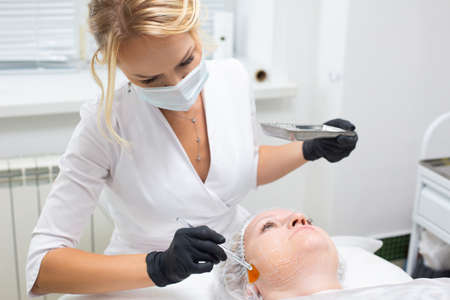 Portrait of a young dermatologist in a medical mask and gloves applying a special gel with a brush to the face of a lying female client before the procedure of non-surgical ultrasound SMAS liftingの写真素材
