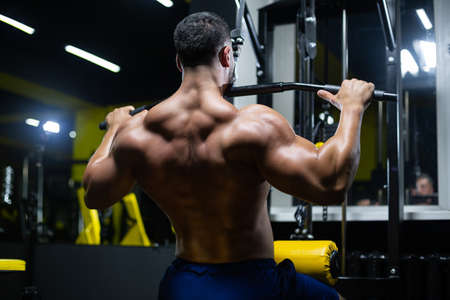Handsome fitness man is performing back workouts using thrust of the upper block machine in a gym,...