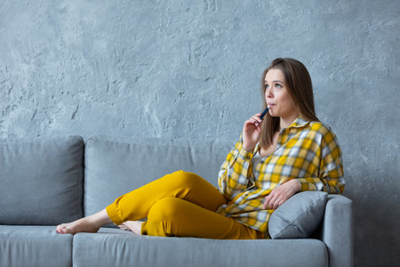 Profile photo of a smiling beautiful girl with an electronic cigarette in her mouth resting on a blue sofa at home.の写真素材