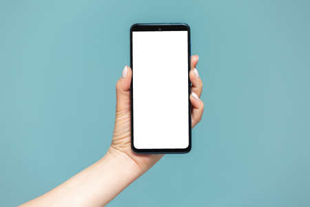 Close-up of a womans hand with a smartphone on which there is a white screen for advertising, on a blue background. Copy paste.の写真素材