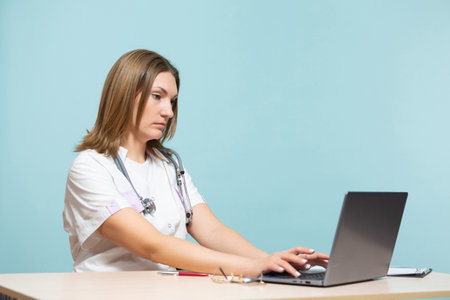 A woman doctor sits at a table and works on a laptop computer, on a blue background. Copy paste. healthcare concept.の写真素材