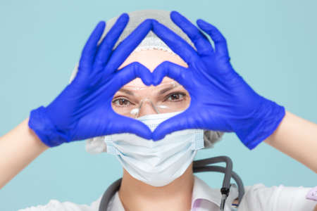 Heart symbol, love. Doctor or nurse woman in a personal protective suit with a stethoscope, on a blue background. In a mask, gloves, goggles and a hat. Copy paste. healthcare concept.の写真素材
