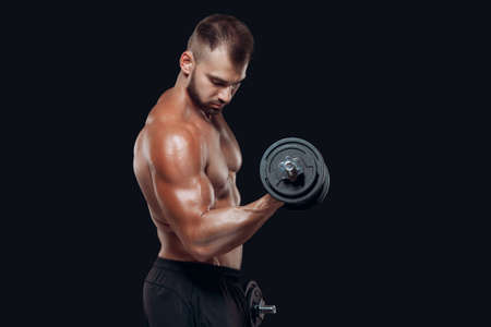Side view of sexy athletic man is showing muscular body with dumbbells, looking at his biceps, isolated over black backgroundの写真素材