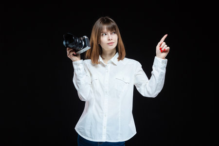Portrait of a young woman wearing a white shirt, holding a camera in her hand and pointing with her hand at your text. The concept of a successful photographer. Isolated black background.の写真素材