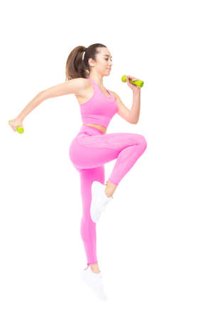 A beautiful, athletic, slim, smiling and cheerful woman in a pink tracksuit performs jumps with dumbbells. Lifestyle concept with sports and gym, moving towards the goal. Isolated on white background.の写真素材