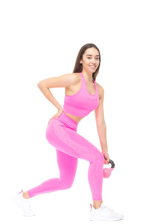 A beautiful, athletic, slim, smiling and cheerful woman in a pink tracksuit performs exercises with a pink kettlebell. Lifestyle concept with sports and gym. Isolated on white background.の写真素材