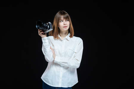 Portrait of a young woman wearing a white shirt holding a camera in her hand. The concept of a successful photographer. Isolated black background.の写真素材