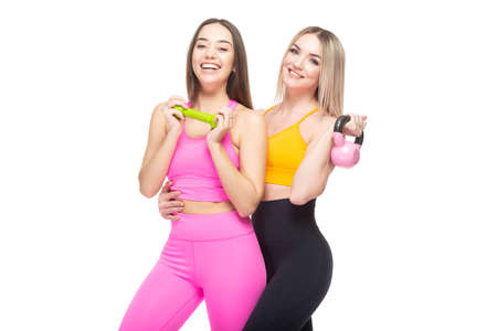 Two beautiful, athletic, slender and cheerful female friends are smiling and holding dumbbells and a kettlebell in their hands. Lifestyle concept with sports and gym. Isolated on white background.の写真素材