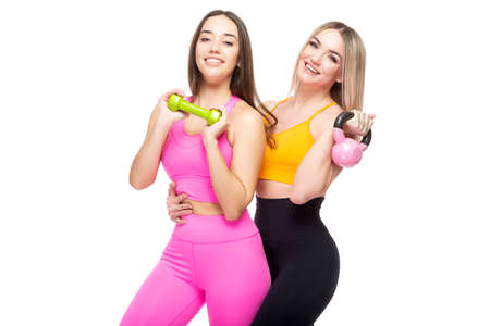 Two beautiful, athletic, slender and cheerful female friends are smiling and holding dumbbells and a kettlebell in their hands. Lifestyle concept with sports and gym. Isolated on white background.の写真素材