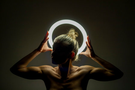 Beautiful young woman with long blond hair and bare back standing with round neon light on dark backgroundの写真素材
