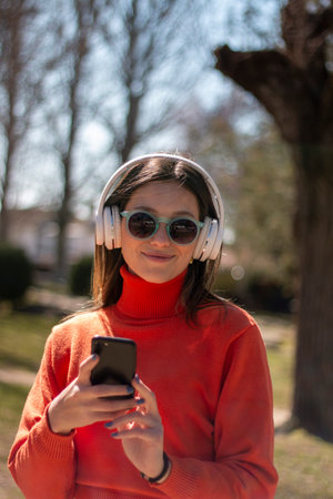 Young brunette woman smiling with headphones, sunglasses and mobile phone in her hand, dressed in orange sweater and jeansの写真素材