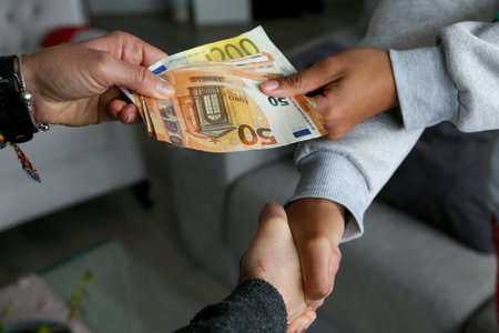 Two women closing a deal, handing over cash and shaking hands; one with light African American skin...