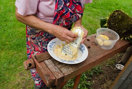 senior woman with apron hands grate peel potatoes with steel shredder tool on wooden outdoor table  