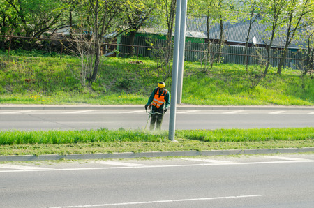 city ??worker with protective mask and vest cuts the grass between the street pavement 