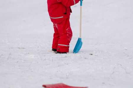child stand with large snow shovel dressed in red waterproof clothing outdoor in winter time の写真素材