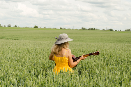 Country girl in yellow dress and hat on head play with guitar at wheat field. の写真素材