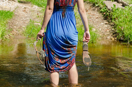 girl holds gray summer sandals and wade barefoot through the fast flowing stream in the park track 