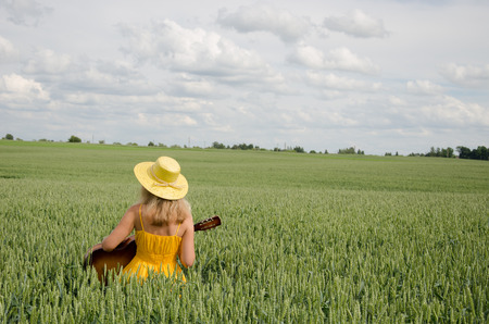 Country woman in yellow dress and hat on head play with guitar at wheat field. の写真素材