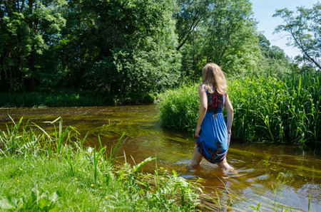 blonde girl with long blue mottled summer dress wade through fast flowing river in summer day の写真素材