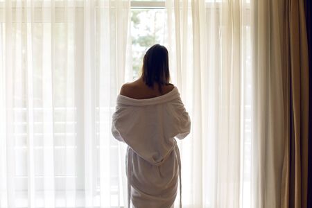 the girl standing at the window his back in a white Terry robe and opens the curtainsの写真素材