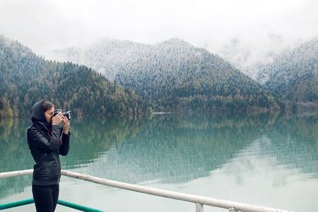 girl in a leather jacket with a camera taking pictures Ritsa lake in the spring morning, with clouds...
