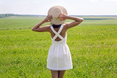 young girl with long dark hair standing on a green field with green grass backs to the camera in a short white summer dress and straw hat and then removes it. throws up his hands and enjoying nature. in the right corner of the screenの写真素材