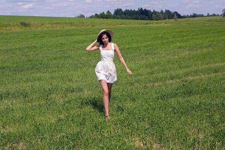 young girl in white dress and holding a straw hat is on the green field with grass and smiles, summer. the model goes on cameraの写真素材