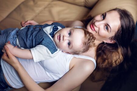 young happy mother in white tank top laying with my son on a light sofa in the apartmentの写真素材