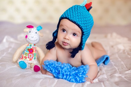 baby is lying on her belly on a big white bed in a big blue hat, and sitting next to a toy goat