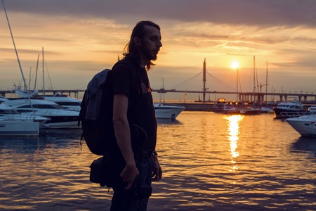 guy with long hair in silhouette standing on the beach with a camera where yachts are docked at the pier at sunset in summer, in the distance, the cable-stayed bridgeの写真素材