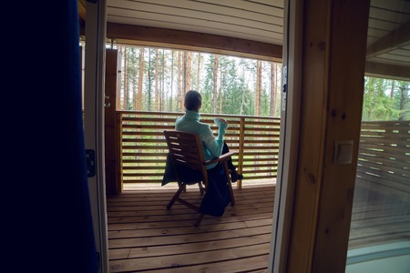 girl in a blue sweater sitting on a wooden chair and drinking tea from a white mug on the veranda of the second floor balcony in the house in the pine forest.の写真素材