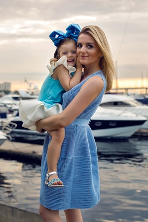 little girl three years sitting on my mother's hands with long blond hair and they are dressed in blue dress and large blue bow on her head , sitting at the pier on the sea with a yacht during sunsetの写真素材