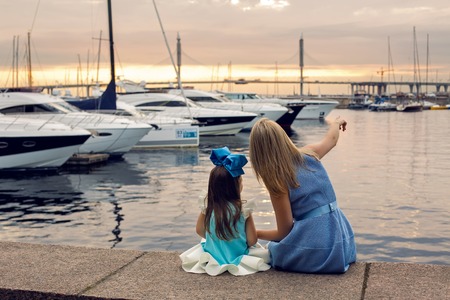 mom shows daughter three years ships in a blue dress and blue big bow sitting on the dock by the sea with yachts at sunsetの写真素材
