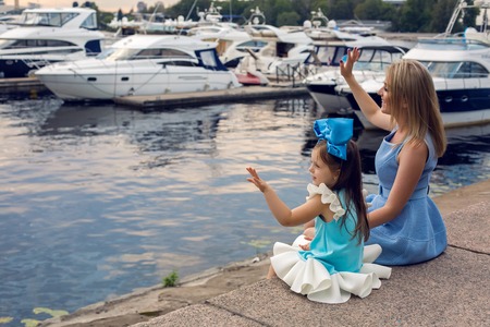 little girl three years sitting with mom with long blond hair and they are dressed in blue dress and large blue bow on her head , sitting at the pier on the sea with a yacht during sunsetの写真素材