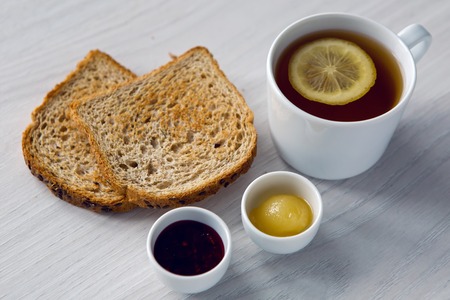 white mug with tea and lemon , jam and honey in small white cups on a white tableの写真素材