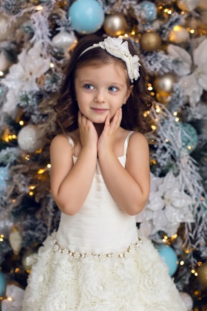 little girl three years old in a white dress smiling at the decorated Christmas tree in roomの写真素材