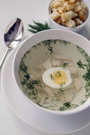 chicken soup in a white plate with a bowl with chicken pieces, croutons and an iron spoon on a white tableの写真素材