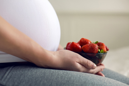 pregnant young girl holding a plate of strawberries sitting in white bedの写真素材