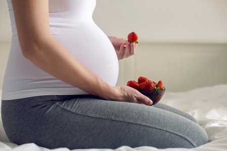 pregnant young girl holding strawberries sitting in white bedの写真素材