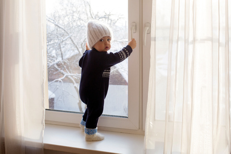 child in a warm blue jumpsuit is on window in winter white knit hatの写真素材