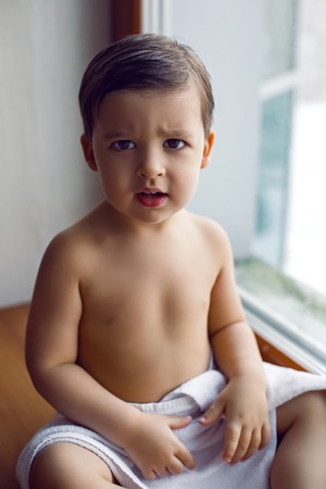 baby boy in bath towel after washing sitting on the windowsill at the big window in winterの写真素材