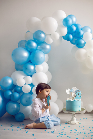 baby boy in blue pants and shirt standing on the floor next to the cake in blue and white balloonsの写真素材
