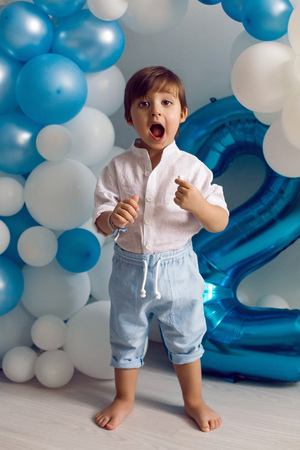 baby boy in blue pants and shirt standing on the floor in blue and white balloonsの写真素材