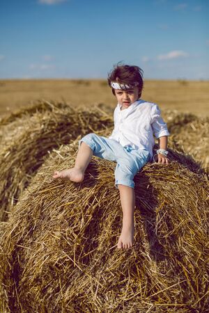 boy child in bandana and white shirt sitting on a bale of hay in the summerの写真素材