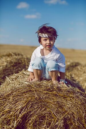 boy child in bandana and white shirt sitting on a bale of hay in the summerの写真素材