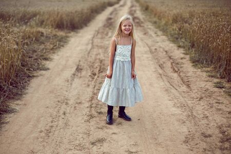 blonde girl stands on an earthen path in a long dress among a wheat field in the summerの写真素材