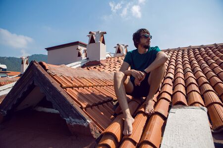 man in the green t-shirt shorts and sunglasses sitting on the roof of a clay orange tileの写真素材